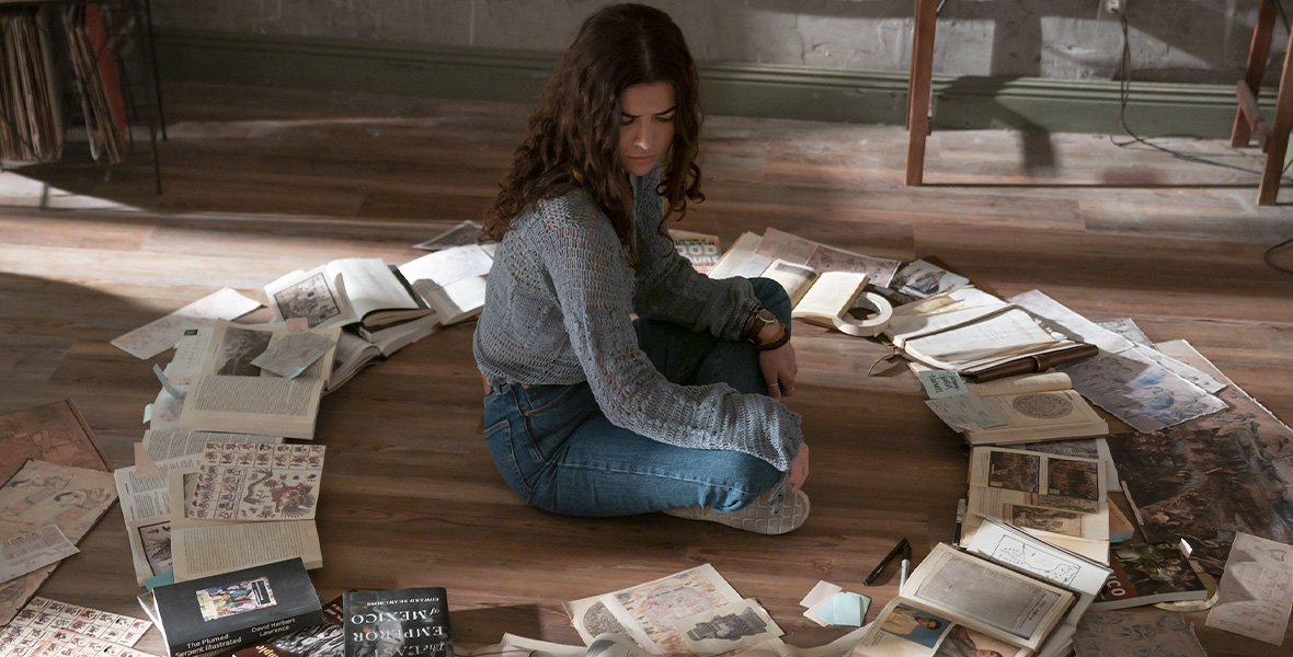 In a scene from Disney+ Original series National Treasure: Edge of History, actor Lisette Olivera portrays Jess and sits cross-legged on the floor. Olivera is surrounded by a circle of books, newspapers, and drawings. Olivera wears a light blue long-sleeved sweater and a pair of light blue denim pants.