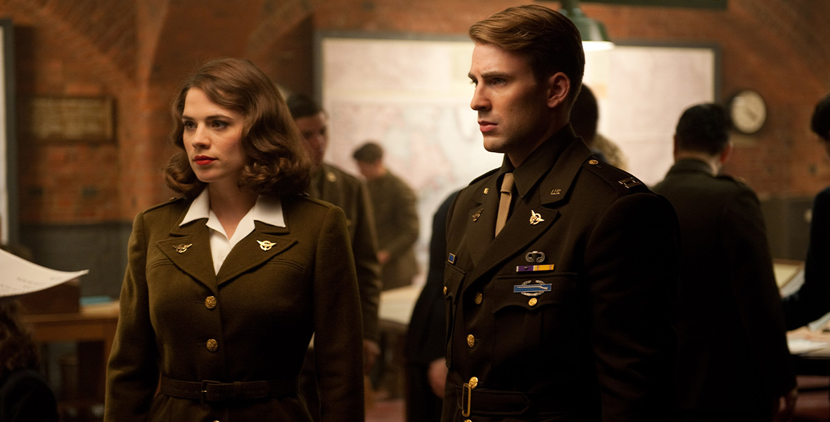 In a scene from Marvel Studios’ Captain America: The First Avenger, actors Hayley Atwell as Peggy Carter and Chris Evans as Steve Rogers/Captain America stand side-by-side and wear olive military uniforms. Behind Atwell and Evans are other soldiers and a large white board with writing and drawings.