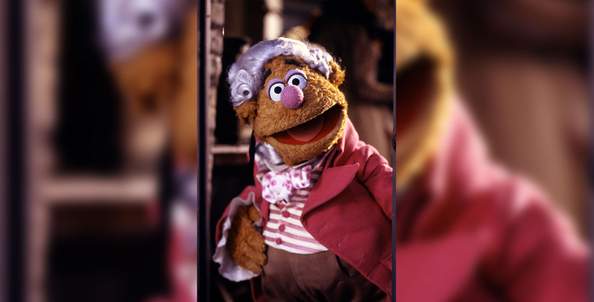 In an image from The Muppet Christmas Carol, Fozzie Bear as Fozziwig is wearing a white wig and a pink coat with a striped pink and a white ascot.