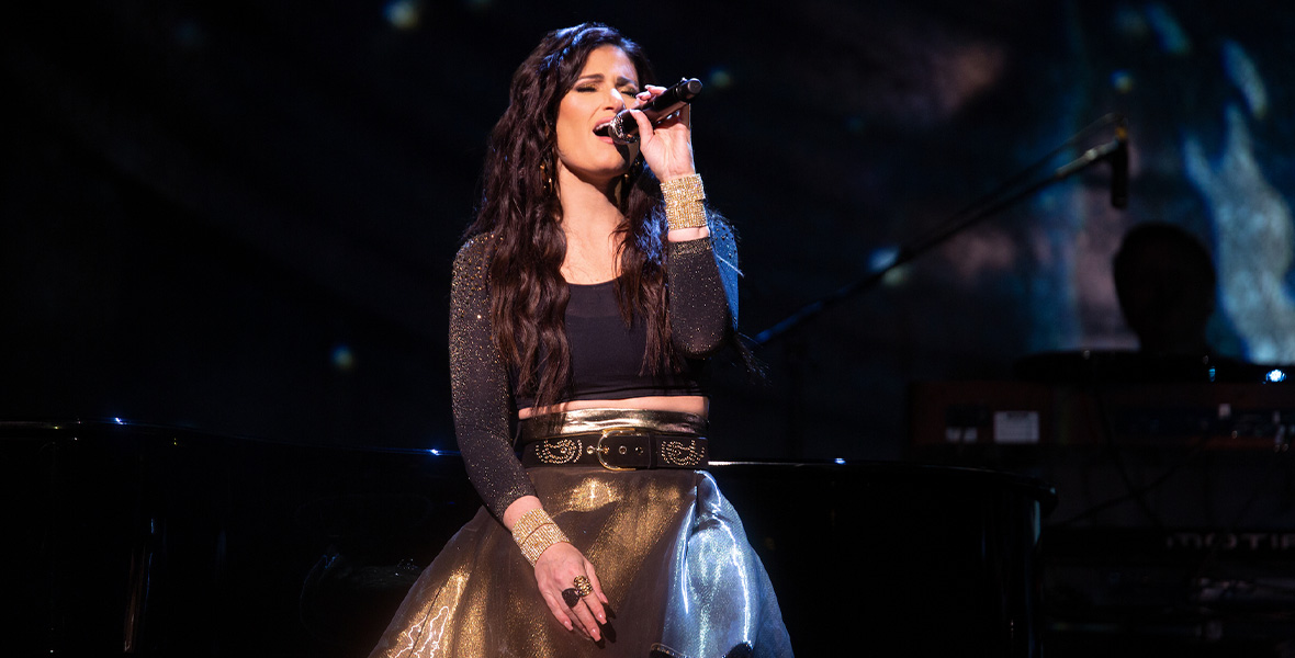In a scene from the Disney+ special Idina Menzel: Which Way to the Stage?, performer and Disney Legend Idina Menzel sits and sings into a black, handheld microphone in her left hand. She wears a black, cropped top with long sleeves adorned with gold glitter, a gold chiffon shirt, black belt, and gold bejeweled bangle bracelets. The stage spotlight shines solely on Menzel, creating a black background behind her.