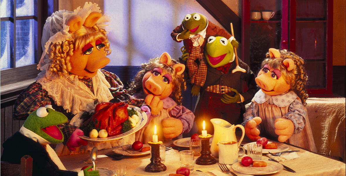 In a scene from Walt Disney Pictures and Jim Henson Productions’ The Muppets Christmas Carol, Muppets Miss Piggy and Kermit the Frog portray character from A Christmas Carol and sit around a small wooden rectangular table. Around Miss Piggy and Kermit the Frog are miniature versions of them playing their children. Miss Piggy wears a white head covering and a red, blue, and white plaid dress with a lace shawl. Miss Piggy holds a white plate with a cooked turkey, greens, and white onions. Kermit the Frog wears a brown suit and brown, orange, and white checkered shirt. On the table are plates, cutlery, and two white candles lit and in brown candle holders.