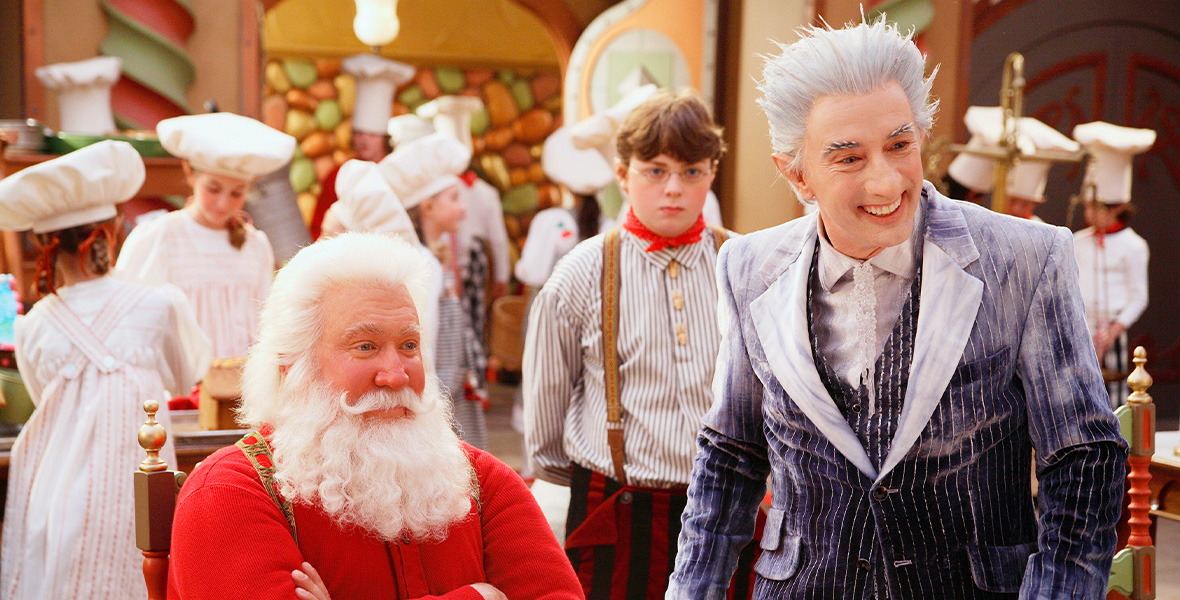 In a scene from The Santa Clause 3: The Escape Clause, actor and Disney Legend Tim Allen portrays Scott Calvin/Santa Claus and sits in a wooden chair inside Santa’s workshop. Allen wears a red, long-sleeved shirt and red and gold suspenders, and a bushy white beard. Actor Martin Short portrays Jack Frost and wears a dark blue three-piece suit with white pinstripes, a white button-down shirt, and an icicle tie. Short’s suit is dusted with a white powdery substance. Behind Allen and Short are children wearing white chef hats, white gowns, and aprons with red pinstripes.