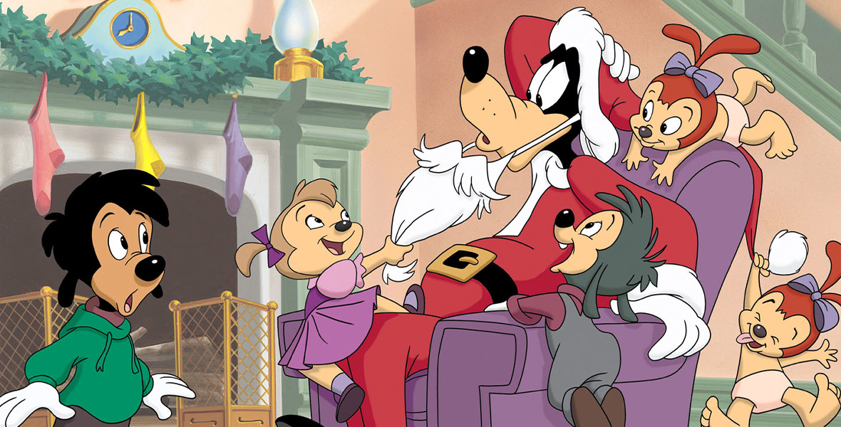 In a scene from Mickey’s Once Upon a Christmas, Goofy, dressed as Santa Claus in a red suit and a long white beard, sits in a purple armchair. Goofy is surrounded by small children who are climbing on the chair and pulling the beard. Max stands to the left and wears a green hooded sweatshirt and a pair of black pants. On the fireplace are a green garland, a lamp, and three colorful stockings.