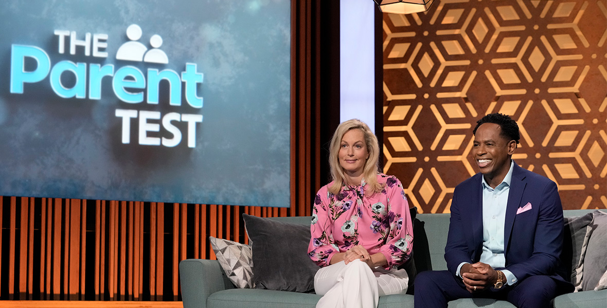 In a scene from ABC series The Parent Test, hosts Ali Wentworth and Dr. Adolph Brown sit side-by-side on a teal couch. Wentworth wears a pair of white pants and a pink blouse with a floral print. Dr. Brown wears a dark blue suit and a light blue button-down shirt. Behind them is a large screen with the series’ title against a soft gray background.