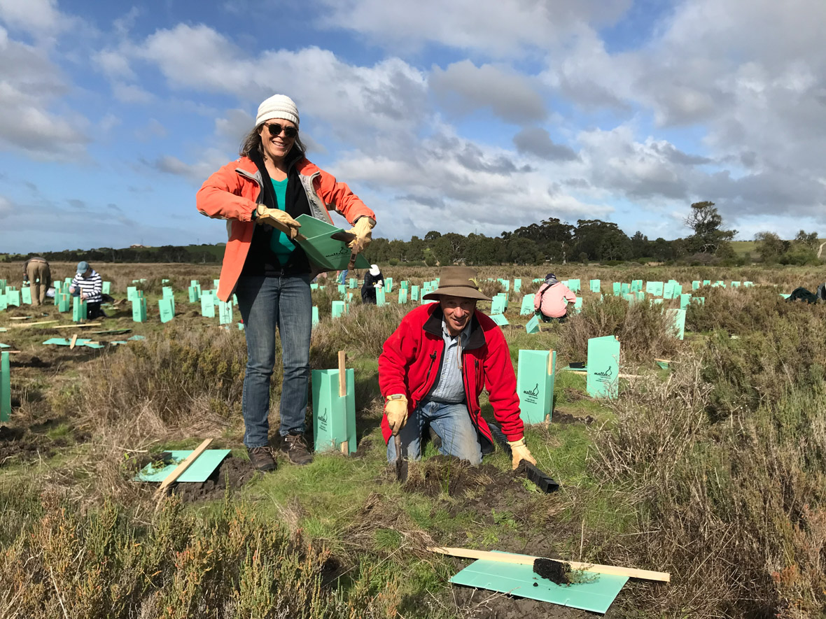 Two people smile for a photo on Watchalunga Planting Day. They are wearing orange and red jackets, respectively, and hats and jeans. They are surrounded by green boxes. All around them, other people are planting Watchalungas.