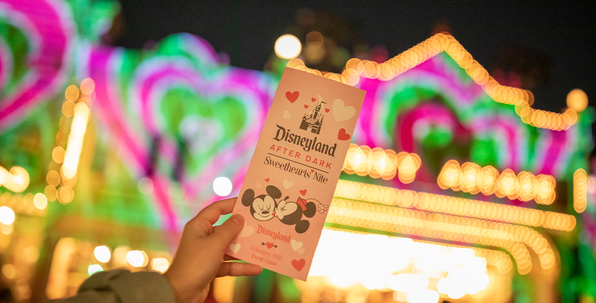 In a promo image for Disneyland After Dark’s Sweethearts’ Nite, an event guide is being held up by a hand, with the bright lights of Disneyland’s Main Street, U.S.A. in the background. Caricatures of Mickey Mouse and Minnie Mouse can be seen on the event guide; Minnie is giving Mickey a kiss on the cheek.
