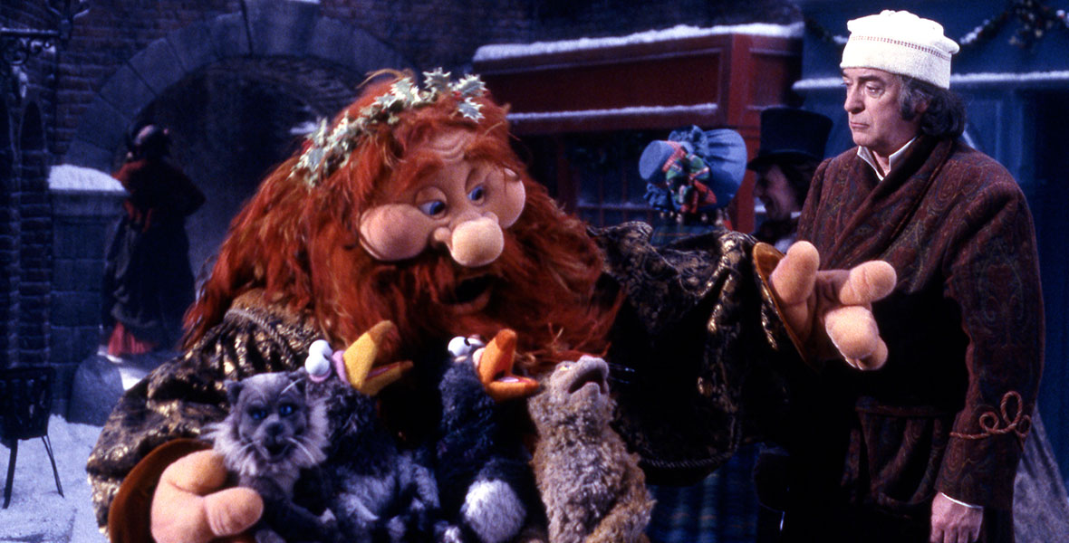 In an image from The Muppet Christmas Carol, the Ghost of Christmas Past—who has long red hair and a long red beard, and wearing a gold brocade robe—is seen on the left, with his arms outstretched around a group of puppet animals including a penguin. To his right, Michael Caine as Ebenezer Scrooge (dressed in his sleeping gown and cap) looks on slightly confused. They are standing in the street; passersby can be seen in the background.