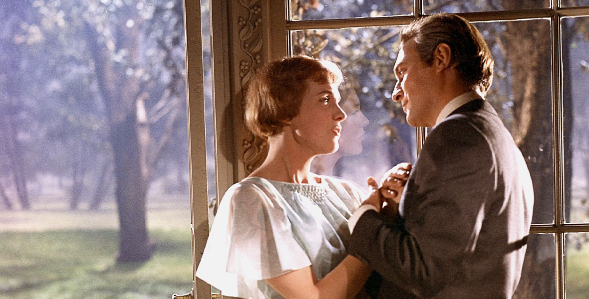 In a scene from The Sound of Music, actor and Disney Legend Julie Andrews stands face to face and locks arms with actor Christopher Plummer. Andrews wears a light blue dress with sheer short sleeves. Plummer wears a dark brown suit and a white button-down shirt. Andrews and Plummer stand next to a large window with intricate carvings.