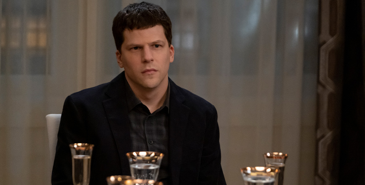 In a scene from FX series Fleishman Is in Trouble, actor Jesse Eisenberg portrays Toby and sits at the end of a dinner table. Eisenberg wears a black button-down shirt and a black blazer jacket. On the table in front of him are wine and champagne glasses, plates, and a piece of chocolate cake.