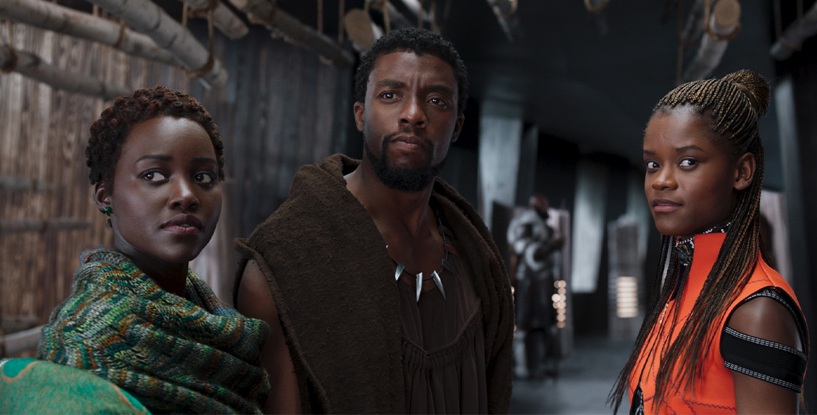 In a scene from Marvel Studios’ Black Panther, actors Lupita Nyong’o as Nakia, Disney Legend Chadwick Boseman as T’Challa/Black Panther, and Letitia Wright as Shuri stand in a narrow hallway. Nyong’o wears a green printed scarf around her neck and looks to her right; Boseman wears a brown hooded top and a metal claw necklace around his neck; Wright wears an orange sleeveless top with black straps on her arms.