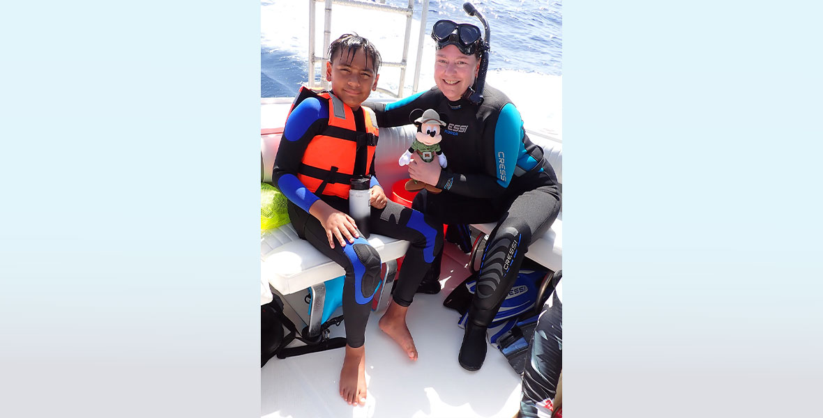 A child in a blue and black wetsuit and an orange and black life vest sits on a white cushion on a boat. Next to him, an adult in a black and blue wetsuit wears a snorkel and holds a Mickey Mouse plush in hand.