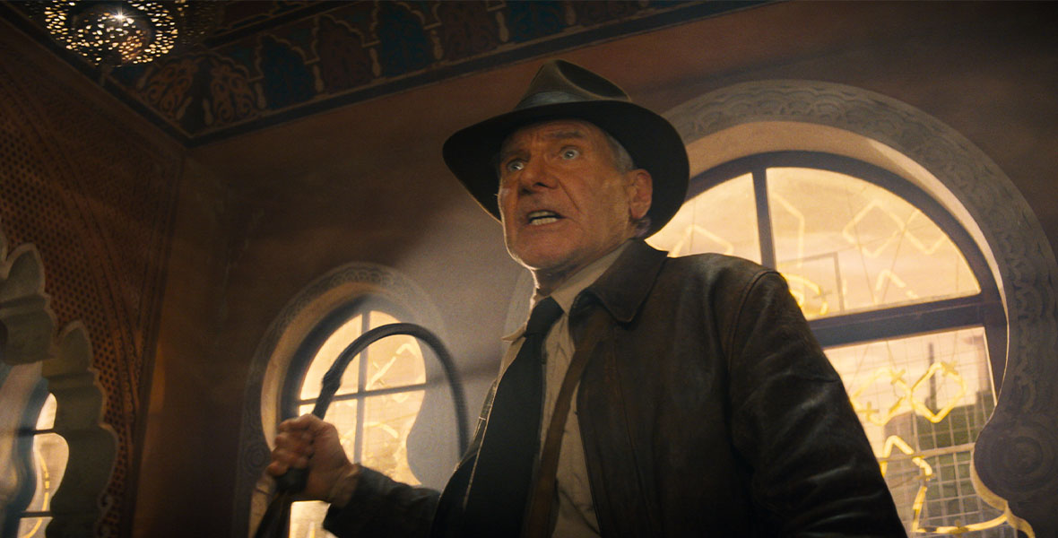 In a scene from Lucasfilm’s Indiana Jones and the Dial of Destiny, actor Harrison Ford portrays Indiana Jones. Ford wears a brown leather jacket, a tan button-down shirt, a brown tie, and a brown fedora. Ford holds a whip in his right hand and stands in front of a large window.