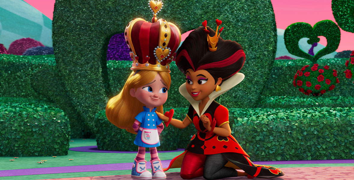 Alice (left), smiling and blonde, wears a blue jacket and a pink apron over a purple and pink striped long-sleeved shirt and matching pants. She wears blue and white high-top shoes with pink trim. Alice wears a tall gold crown with red and maroon fabric, adorned with diamonds, rubies, and red hearts. She stands with her hands behind her back. The Queen of Hearts rests her right hand on Alice’s shoulder. The Queen of Hearts wears a smaller gold grown on her head, with red hearts on the tip of each prong. Her black bouffant has two red streaks on each side. She wears a black and red peplum top with an exaggerated white collar, as well as red and black pants. She has a red bracelet, in the shape of a heart, on each wrist. She is kneeling on a rug.
