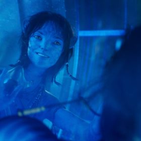 Kiri, a Na’vi teen with blue skin, presses her hands to a pane of glass and smiles. She wears a beaded necklace and a braided armband.