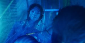 Kiri, a Na’vi teen with blue skin, presses her hands to a pane of glass and smiles. She wears a beaded necklace and a braided armband.