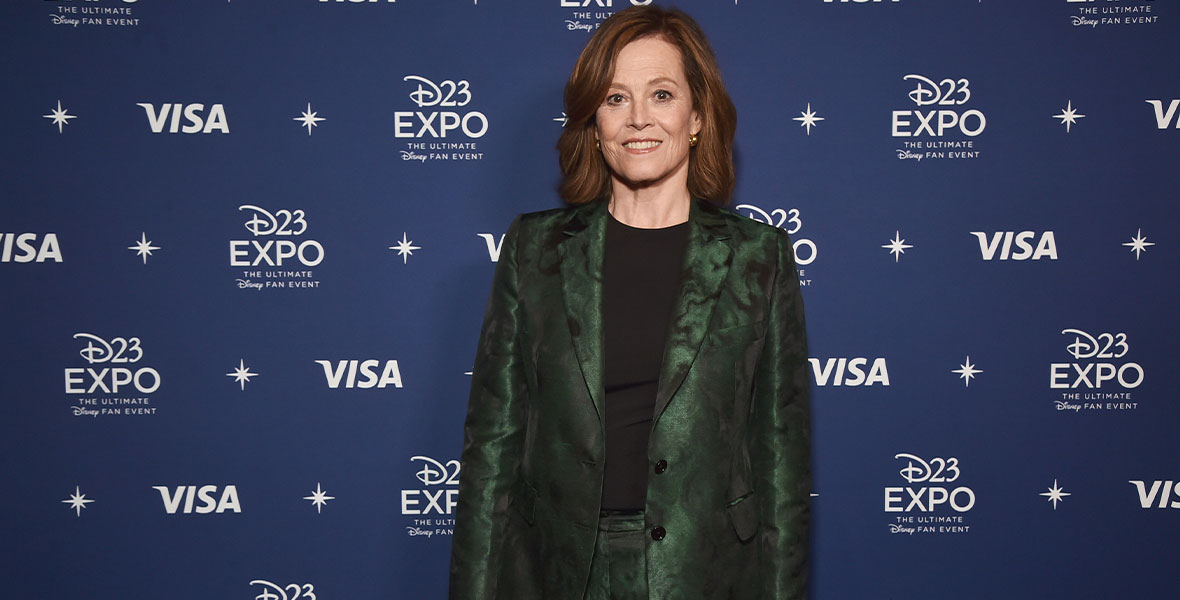 Sigourney Weaver smiles while posing for photos against a D23 Expo backdrop. She wears a dark green suit and a black T-shirt.