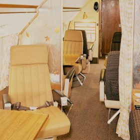 A photo from the Walt Disney Archives of the interior of Walt Disney’s plane, affectionately known as “The Mouse,” as it appeared in the 1960s. A single seat faces a small table on the left side, while bench seats face the table on the right. A yellow-ish, semi-transparent screen with autumn leaves on it divides this section of the plane from the rows of seats behind it.