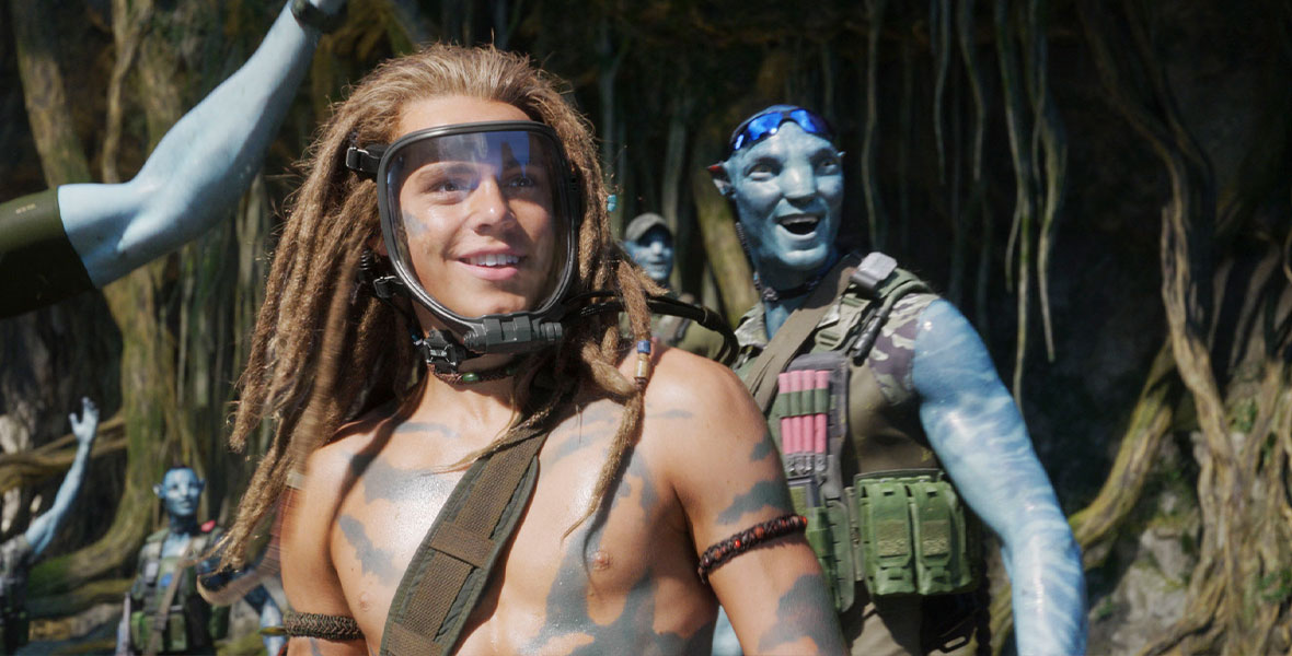 Spider, played by Jack Champion, wears an exopack—a clear mask with a black frame that helps him breathe—over his face. His light brown hair is in dreadlocks. He is shirtless and surrounded by Na’vi, a species of blue, extraterrestrial humanoids.
