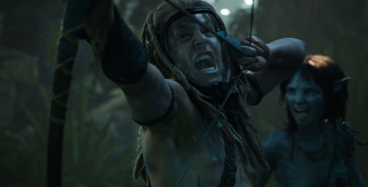 Spider, played by Jack Champion, is shirtless and wears an exopack—a clear mask with a black frame that helps him breathe—over his face. His light brown hair is in dreadlocks. He wields a bow and arrow and is surrounded by a lush green forest. Behind him is Kiri, a blue, 14-year-old member of the Na’Vi clan.