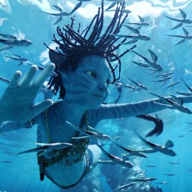 The blue Na’vi child Tuktirey, aka Tuk, swims underwater among other marine life. Her many braids float upward, and her four fingers on each hand are outstretched as she surveys the area around her.