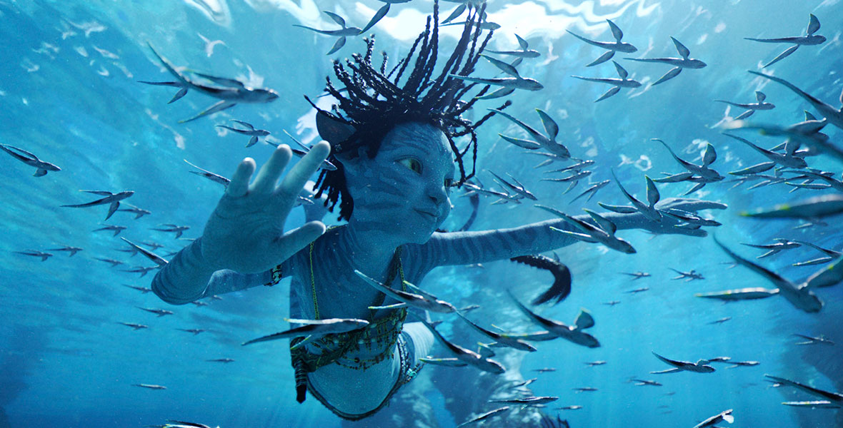 The blue Na’vi child Tuktirey, aka Tuk, swims underwater among other marine life. Her many braids float upward, and her four fingers on each hand are outstretched as she surveys the area around her.