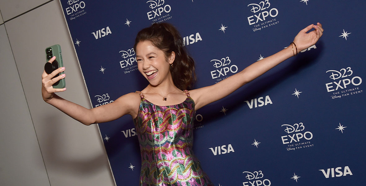 Trinity Bliss wears a metallic rainbow mini dress with spaghetti straps. Her curled hair is in a high ponytail, and she has both arms extended. In her right hand, she holds a green iPhone and snaps a selfie. Behind her is a D23 Expo step and repeat.
