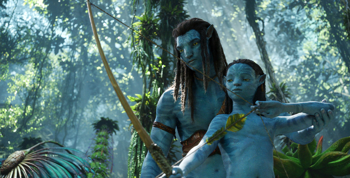 The blue Na’vi Jake (left) stands behind his son, Neteyam (right), who wields a bow and arrow in a dense, lush forest.