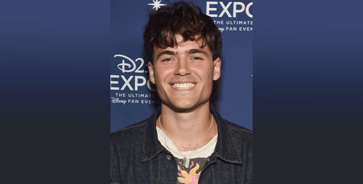Jamie Flatters wears a denim jacket, a graphic T-shirt, and layered silver necklaces. His brown, curly hair is messy, and he is smiling wide. Behind him is a D23 Expo step and repeat.