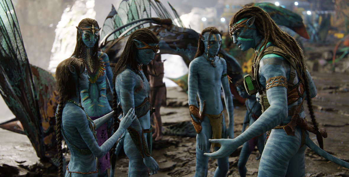 Members of the Sully family—Kiri (left, front), Neytiri (left, back), Neteyam (center), Lo’ak (right, back), and Jake (right, front)—stand in a circle. Neteyam has his head down, and Kiri puts her arms on his back and arm to console him. Jake is speaking to his family, with his arms outstretched, as Neytiri and Lo’ak listen intently.