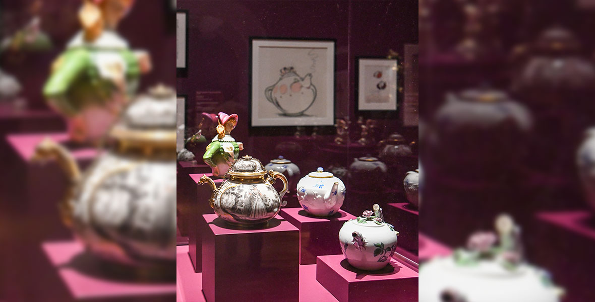 Rococo teapots with intricate designs and anthropormorphistic features are displayed in a gallery case in front of Disney animation art of Mrs. Potts from Beauty and the Beast at the exhibition Inspiring Walt Disney: The Animation of French Decorative Arts on view at The Huntington.