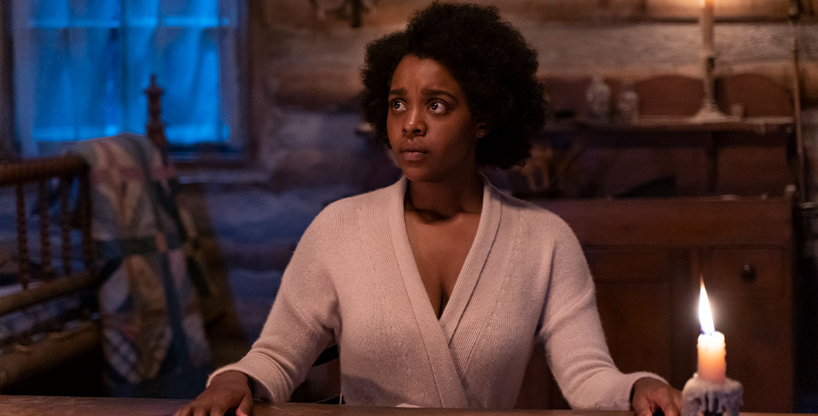 In a scene from the FX series Kindred, actor Mallori Johnson portrays Dana James and sits at a wooden table with each hand resting on the top. Johnson looks to her left with wide, teary eyes. Johnson wears a tan, long-sleeved sweater. On the table is a single metal candle holder covered in wax. Johnson sits inside a dimly lit wooden cabin. Behind Johnson are an old metal candelabra on the wall and a wooden rocking chair with a multi-colored quilt draped on the back of the chair.
