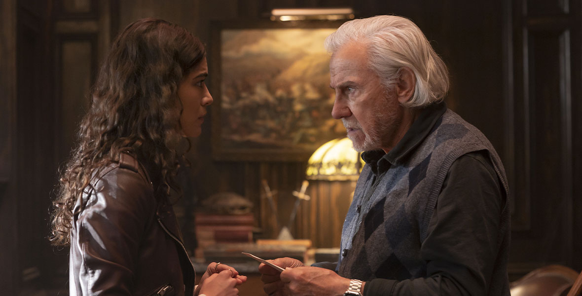In a scene from the Disney+ Original series National Treasure: Edge of History, from left to right, actors Lisette Olivera as Jess Valenzuela and Harvey Keitel as Peter Sandusky stand face-to-face inside a dimly lit room covered with wood paneling. Olivera wears a brown leather jacket on top of a long-sleeved cream top. Keitel wears a charcoal long-sleeved top, a charcoal sweater vest with a black argyle design, and a silver watch on his left wrist. Behind Olivera and Keitel are a large wooden bureau—topped with old books, an antique glass lamp, and two daggers in a jar—and a large landscape painting with an ornate golden frame.