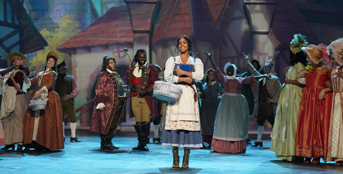 In a scene from the ABC special Beauty and the Beast: A 30th Celebration, musical performer HER portrays Belle, smiles, and stands center. HER holds a book with a royal blue book jacket against her chest. A white wicker basket dangles on her right arm. H.E.R. wears a white blouse, a blue jumper, and a white apron adorned with several red flowers, and brown leather booties. Behind HER are a crowd of townspeople dressed in colorful period ensembles and a large backdrop painted to resemble a small town with cream buildings and red-tiled roofs.