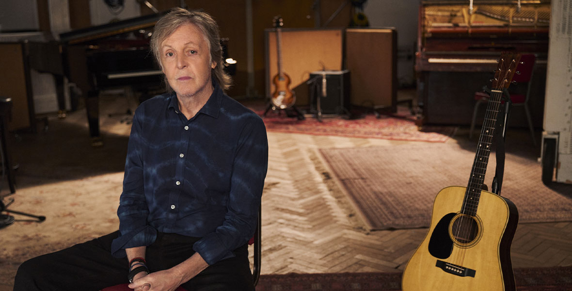 In a scene from the Disney Original Documentary If These Walls Could Sing, musician Paul McCartney sits in a red-padded chair inside a recording studio. McCartney wears a dark blue button-down shirt with an abstract light blue design and a pair of black pants. Surrounding McCartney are a piano, guitars, an organ, amps, speakers, and large microphones.