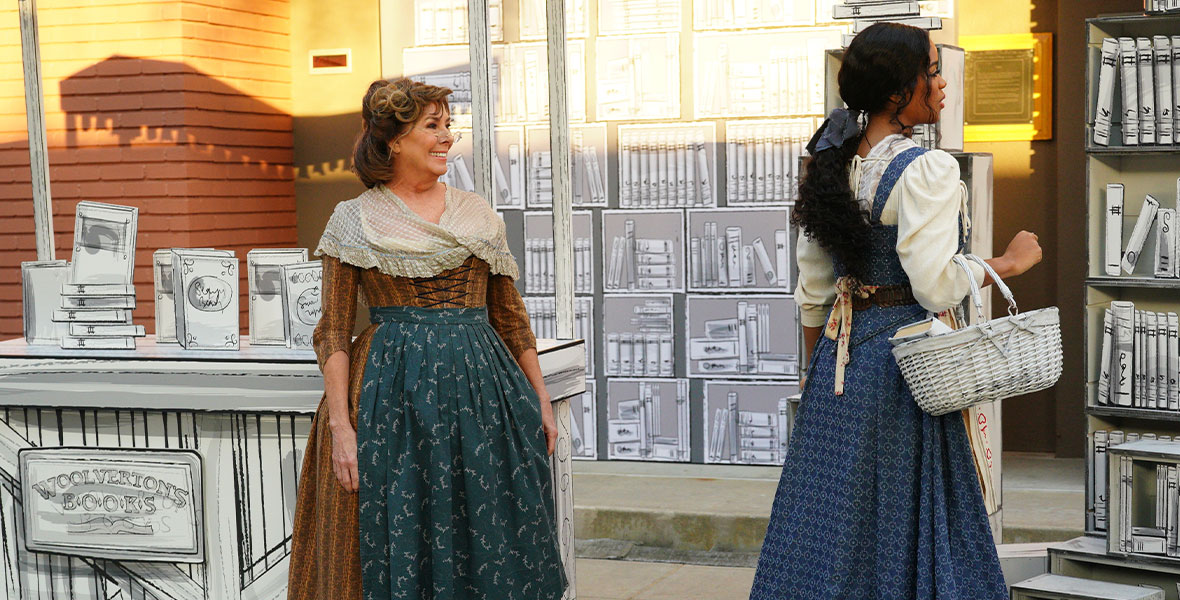 In a still from ABC’s Beauty and the Beast: A 30th Celebration, Paige O’Hara, wearing a brown and orange striped dress, a blue and white apron, and a white shawl, stands in front of Woolverton’s Books. H.E.R., wearing a blue dress, a white shirt, and a blue bow in her hair, peruses the book selection. She carries a white wicker basket in her arm.