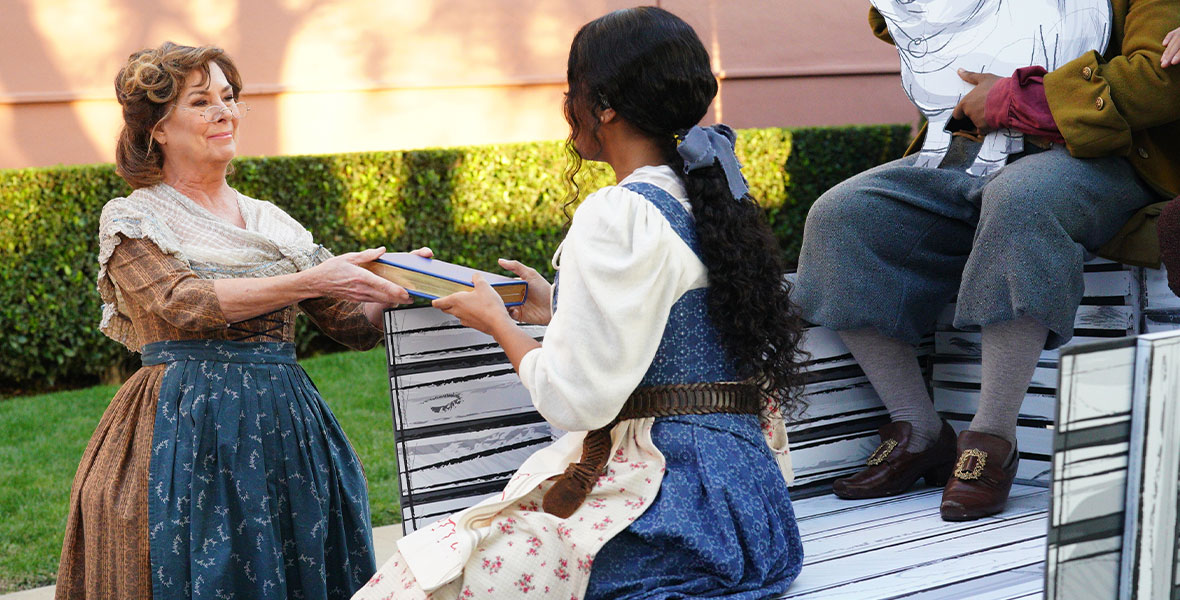 In a still from ABC’s Beauty and the Beast: A 30th Celebration, Paige O’Hara, wearing a brown and orange striped dress, a blue and white apron, and a white shawl, hands a book to H.E.R., wearing a blue dress, a white shirt, and a blue bow in her hair. Behind them, a man caresses an illustration of a pig.