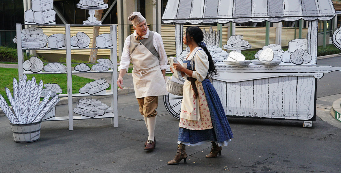 In a still from ABC’s Beauty and the Beast: A 30th Celebration, Richard White, wearing an apron, a chef’s hat, bloomers, and brown buckled shoes, walks with H.E.R., wearing a blue dress, a white shirt, and a blue bow in her hair.