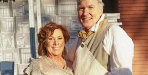 Paige O’Hara, wearing a brown and orange striped dress, a blue and white apron, and a white shawl, poses for a photo with Richard White, wearing an apron, a chef’s hat, and bloomers.