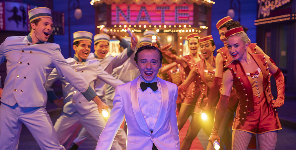 Rueby Wood stars as Nate in 20th Century Studios’ Better Nate Than Ever Wood wears a white suit jacket, a white shirt, black pants, and a black bowtie. He is standing center stage. Behind him are male dancers on one side and female dancers on the other, as well as a marquee that says NATE in block letters.