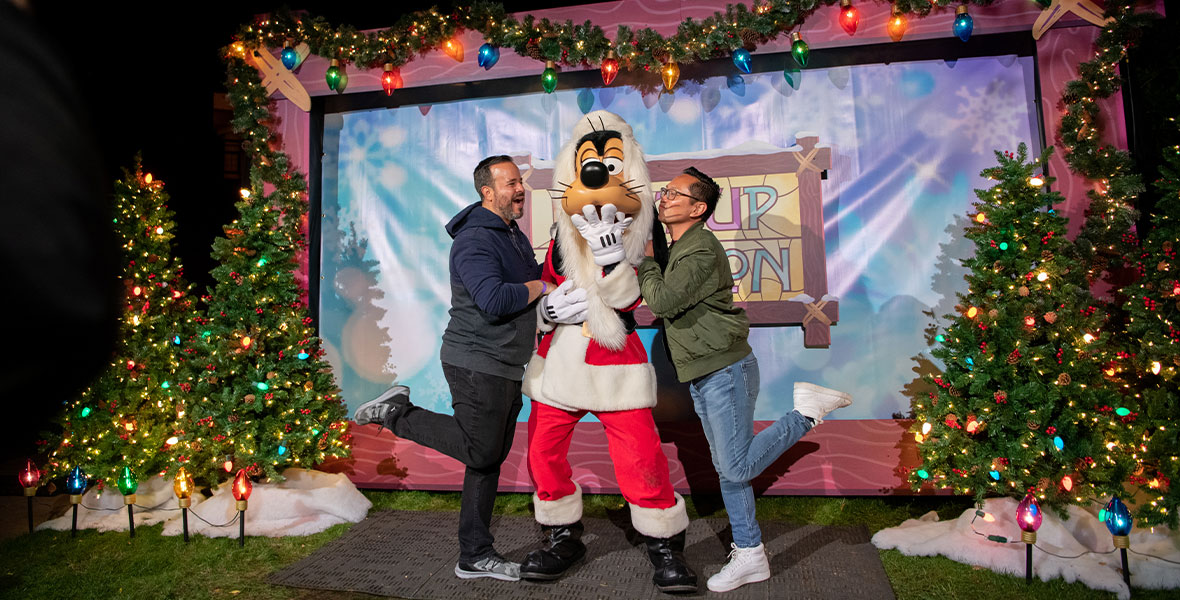 Two guests posing with Santa Goofy in front of a Light Up the Season photo backdrop. The backdrop is blue and snowy with the Light Up The Season logo of stained glass in yellow, blue and pink framed by wood in the center of the backdrop. The guest on the left of Santa Goofy is holding Goofy’s arm with their foot popped up. The guest is wearing a grey and blue hoody with gray jeans. Santa Goofy is in the middle with his hand covering his mouth in excitement. Santa Goofy has a red santa hat, long white beard, and a red and white Santa suit. The guest to the right of Goofy is holding Goofy’s arm with their foot popped up and kissing Goofy’s cheek. The guest is wearing a green jacket and blue jeans.