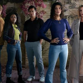 From left to right, Antonio Cipriano, Jake Austin Walker, Zuri Reed, Jordan Rodrigues, Lisette Olivera, Catherine Zeta-Jones, and Lyndon Smith pose for a National Treasure: Edge of History cast photo. They are all wearing streetwear. Behind them is a brick wall, with overgrown purple and green plants cascading down the wall.