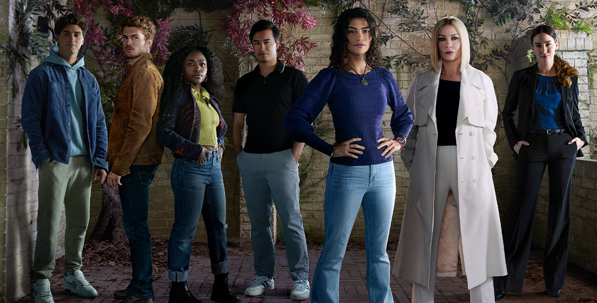 From left to right, Antonio Cipriano, Jake Austin Walker, Zuri Reed, Jordan Rodrigues, Lisette Olivera, Catherine Zeta-Jones, and Lyndon Smith pose for a National Treasure: Edge of History cast photo. They are all wearing streetwear. Behind them is a brick wall, with overgrown purple and green plants cascading down the wall.