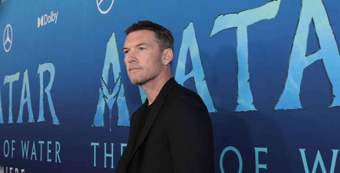 A three-quarters view of Sam Worthington, standing in front of the Avatar: The Way of Water step and repeat