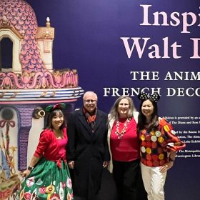Four guest standing in front of Inspiring Walt Disney exhibit logo poster. The poster is purple with a pink and white vase on the left and Inspiring Walt Disney: The Animation of French Decorative Arts on the right. The guest on the left is wearing a red sweater and green holiday skirt. The guest to the right is wearing a black jacket, black pants, and red scarf. The guest to the right is wearing a red shirt, white sweater, and black pants. The guest to the right is wearing a red sweater and red pants.