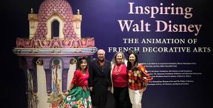 Four guest standing in front of Inspiring Walt Disney exhibit logo poster. The poster is purple with a pink and white vase on the left and Inspiring Walt Disney: The Animation of French Decorative Arts on the right. The guest on the left is wearing a red sweater and green holiday skirt. The guest to the right is wearing a black jacket, black pants, and red scarf. The guest to the right is wearing a red shirt, white sweater, and black pants. The guest to the right is wearing a red sweater and red pants.