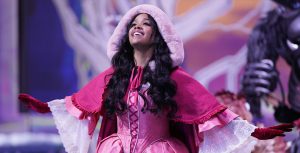 HER wears a pink, fur-lined coat and a pink dress. Her arms are outstretched, and she is smiling wide.