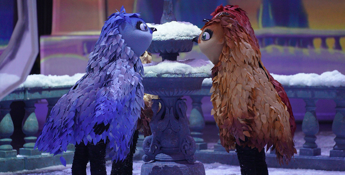 Dancers dressed as a blue bird and a brown bird stare at each other. Behind them is a snow-covered fountain.