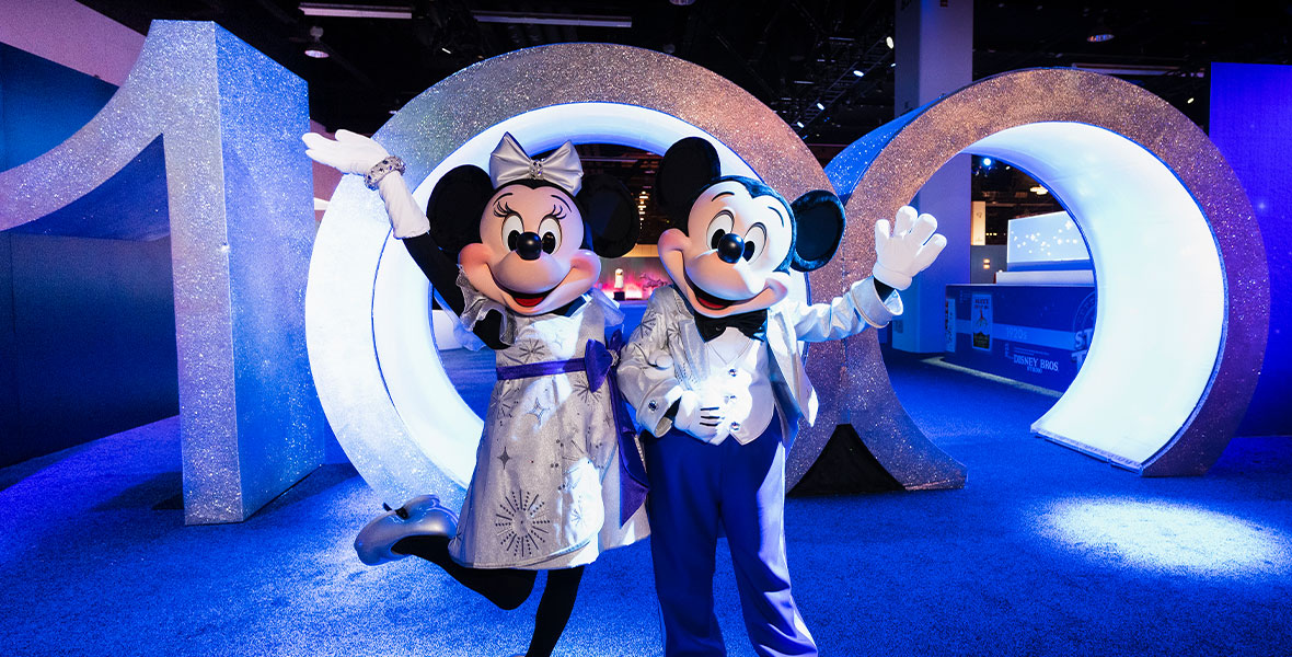 Minnie Mouse, on the left, and Mickey Mouse, on the right, strike a celebratory pose in their Disney100 platinum-colored outfits. They are standing in front of a giant walk-through rendition of the numeral 100, which served as the entryway to the Walt Disney Archives pavilion at the D23 Expo in September in Anaheim, California.