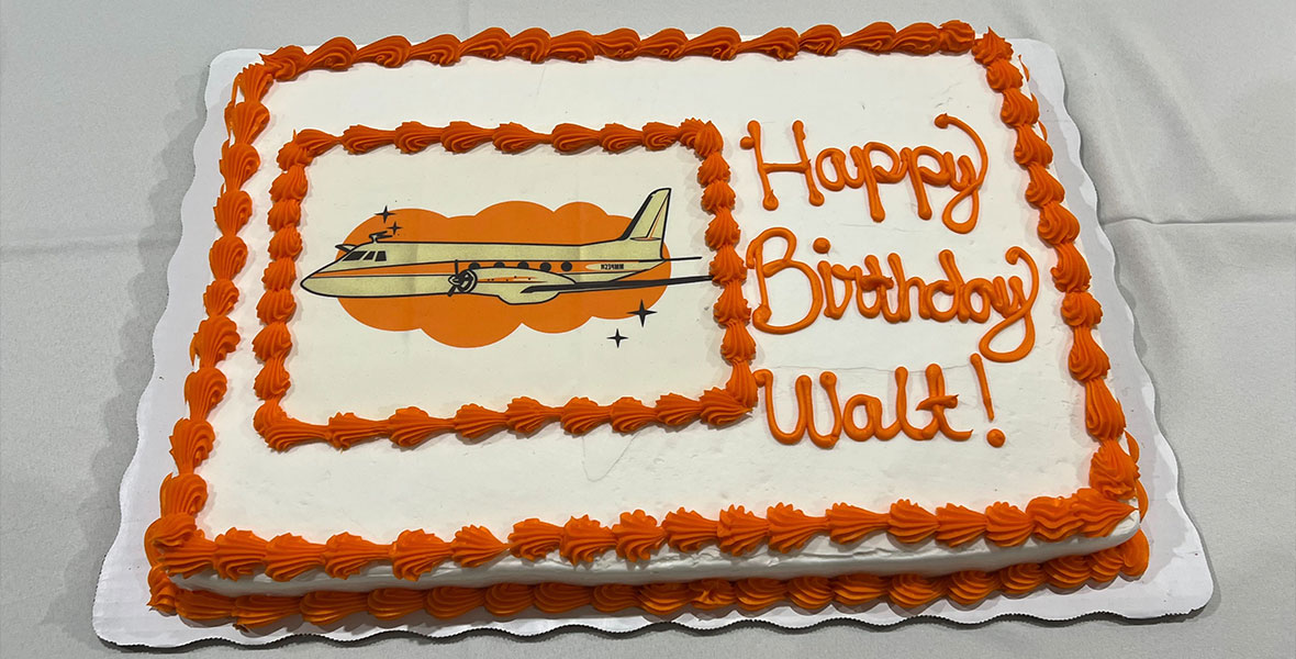 Walt Disney’s Birthday cake with white and orange frosting. On the left of the cake is a decoration of Walt’s Plane. To the right of the decoration reads Happy Birthday Walt in orange frosting.