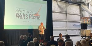 Michael Vargo on stage welcoming guests to the event. Vargo is wearing a gray blazer over a white shirt with black slacks. Vargo is standing in front of a screen displaying the event logo. The room is filled with guests sitting at circular tables watching Vargo.